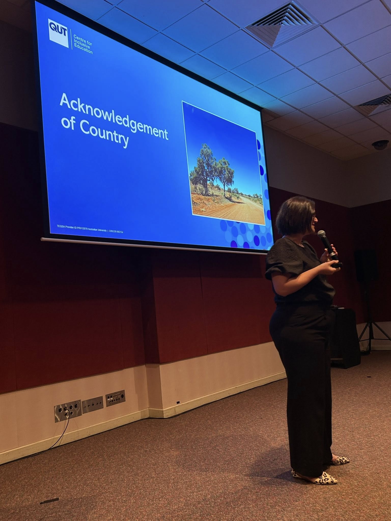 Dr Haley Tancredi gives and Acknowledgement of Country while standing in front of a slide show holding a microphone.
